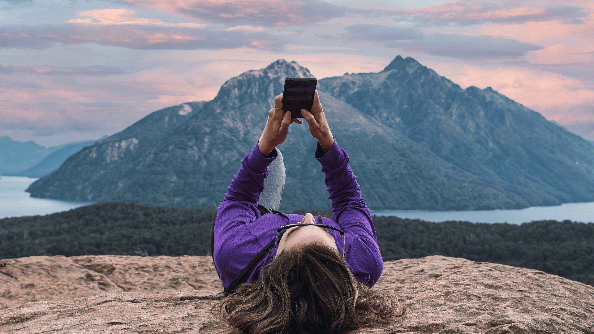 woman checking phone while laying down on a hike with mountain in the background